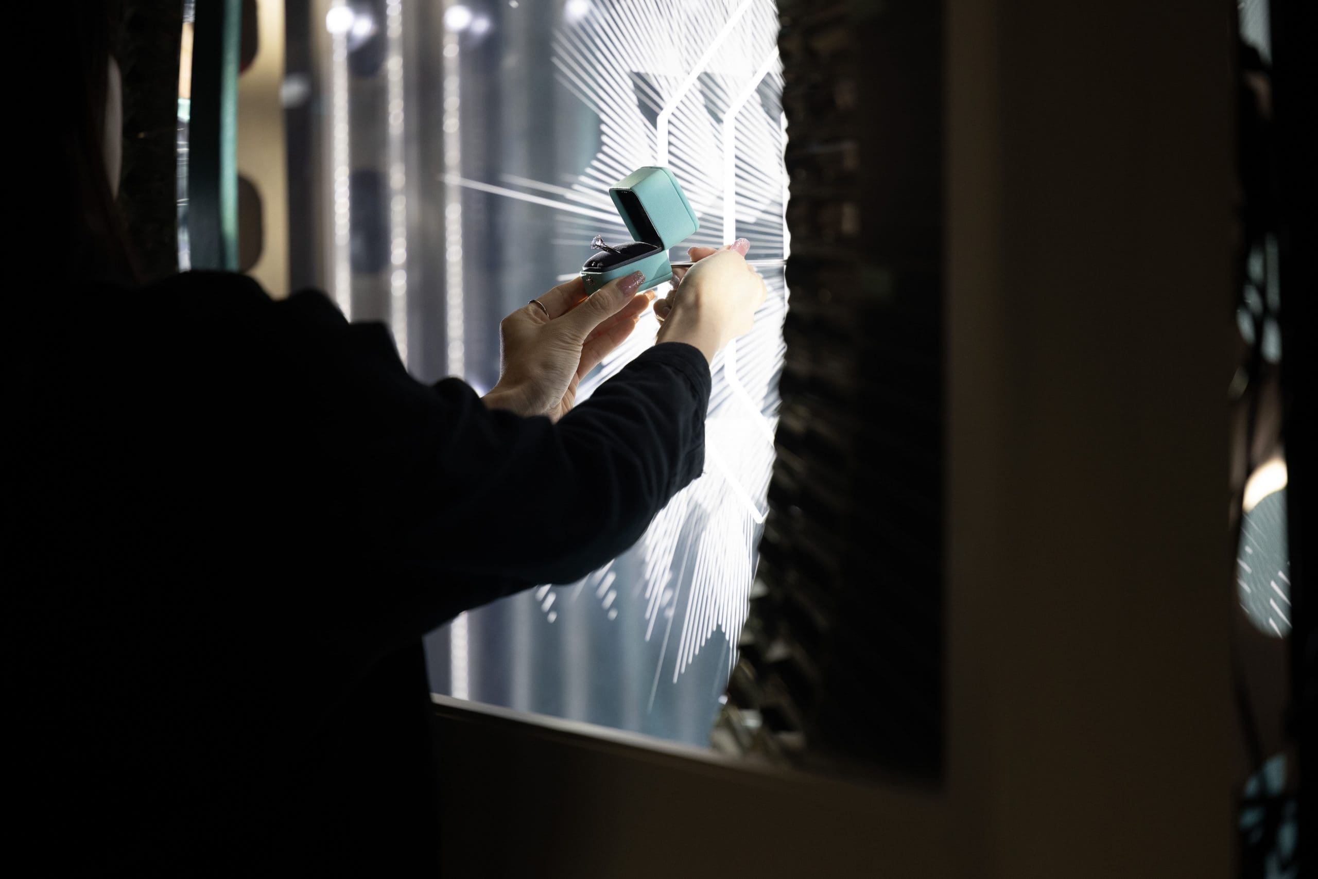 RIOT, NYC Creative Agency: Artisan placing a sparkling Tiffany ring in its box in front of a backlit geometric window display, showcasing intricate light patterns Artisan placing a sparkling Tiffany ring in its box in front of a backlit geometric window display, showcasing intricate light patterns