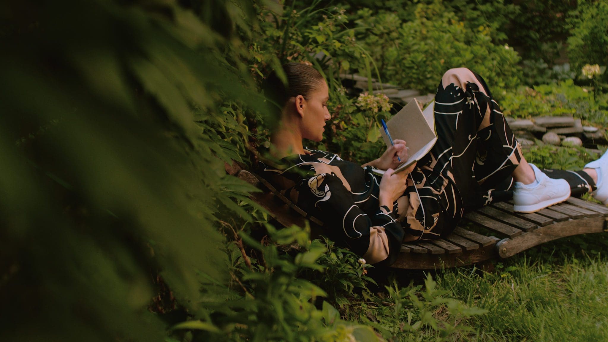 Niia relaxing in a lush garden while writing, wearing Reebok Classic Leather shoes – Take A Walk Series.