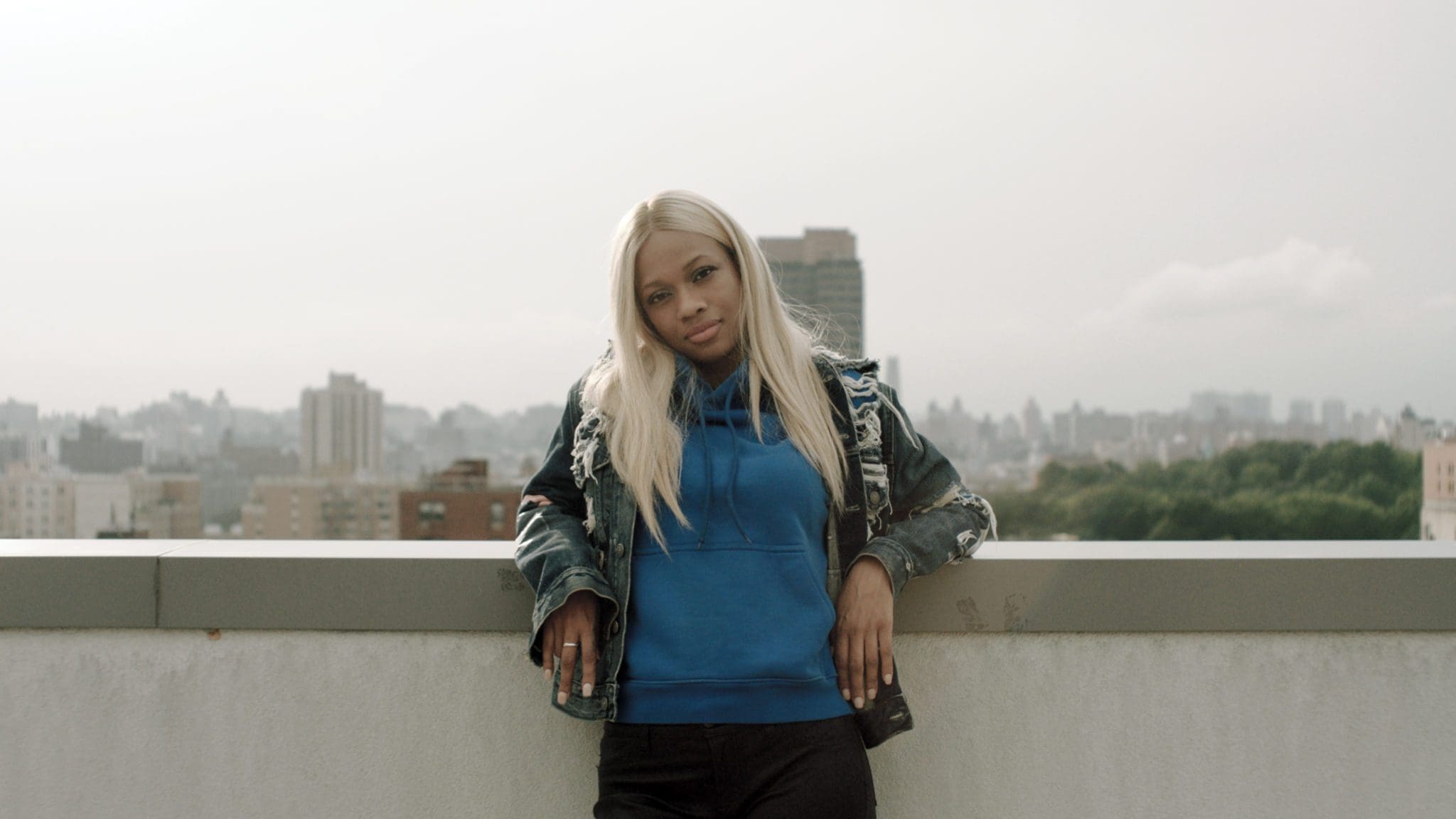 Fatima B standing on a rooftop in New York City, wearing a blue hoodie and denim jacket, from the Reebok Take A Walk series.