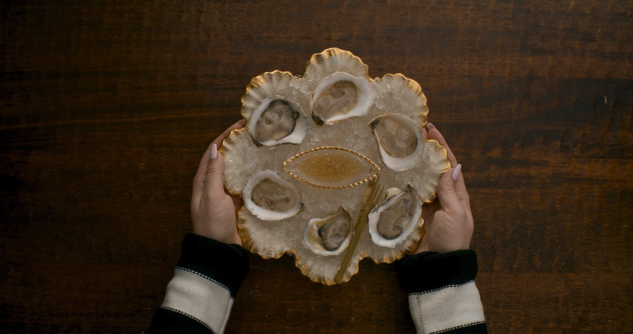A beautifully presented platter of oysters on ice, held by hands, with golden accents and a vintage feel, showcasing Jamie Malone's culinary artistry.