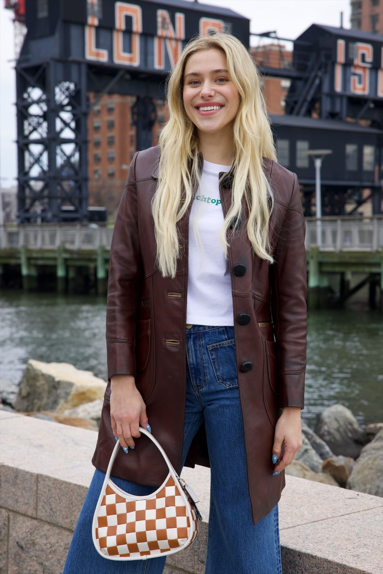 Natalia Spotts in a brown leather coat with a checkered bag on the waterfront