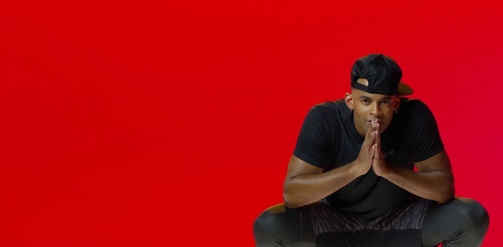 RIOT, NYC Creative Agency: Mike Press crouching with hands clasped, wearing a backward cap, against a striking red background. Mike Press crouching with hands clasped, wearing a backward cap, against a striking red background.