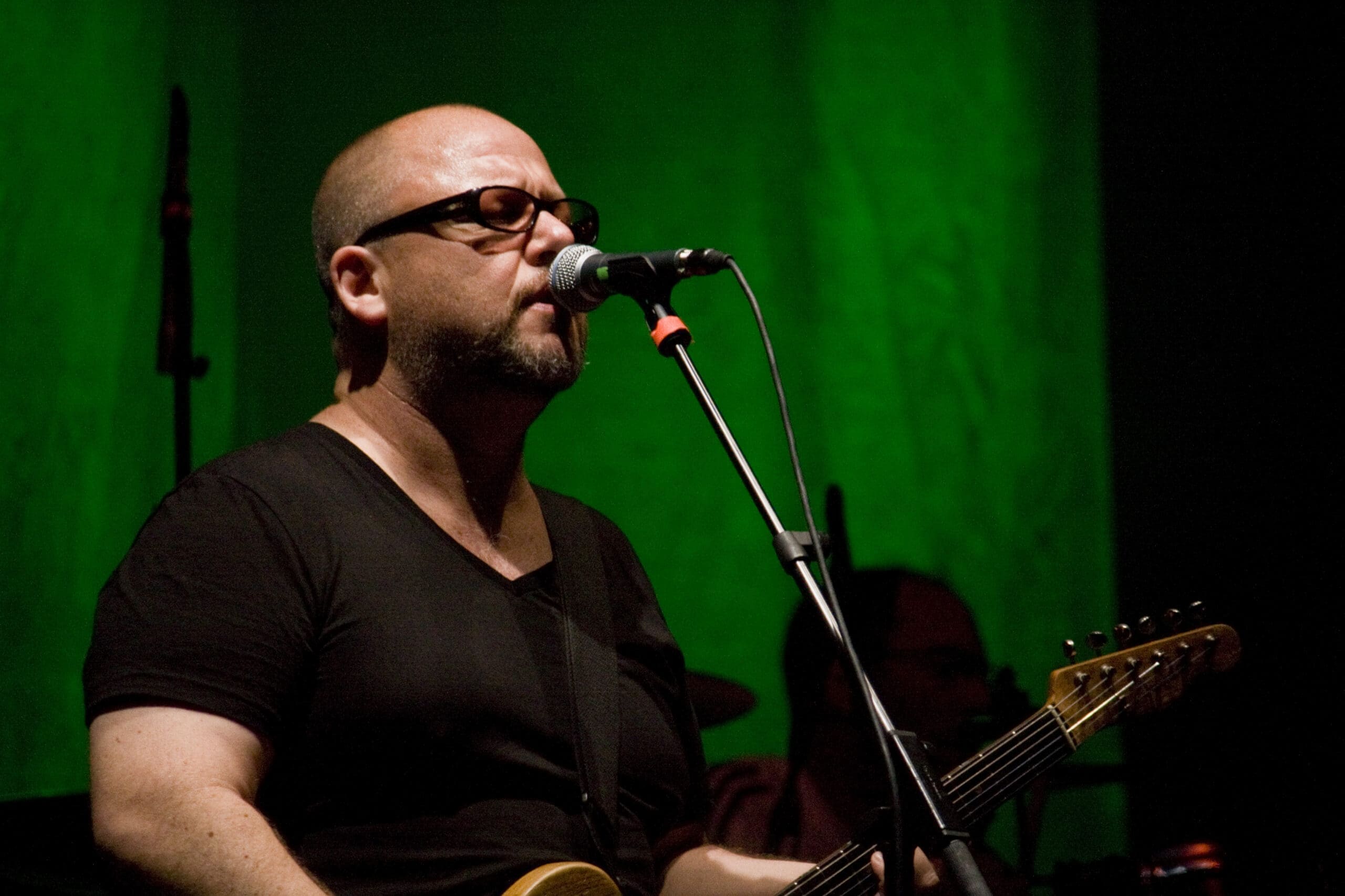 RIOT, NYC Creative Agency: rank Black of the Pixies performing live, singing into a microphone while playing guitar. rank Black of the Pixies performing live, singing into a microphone while playing guitar.