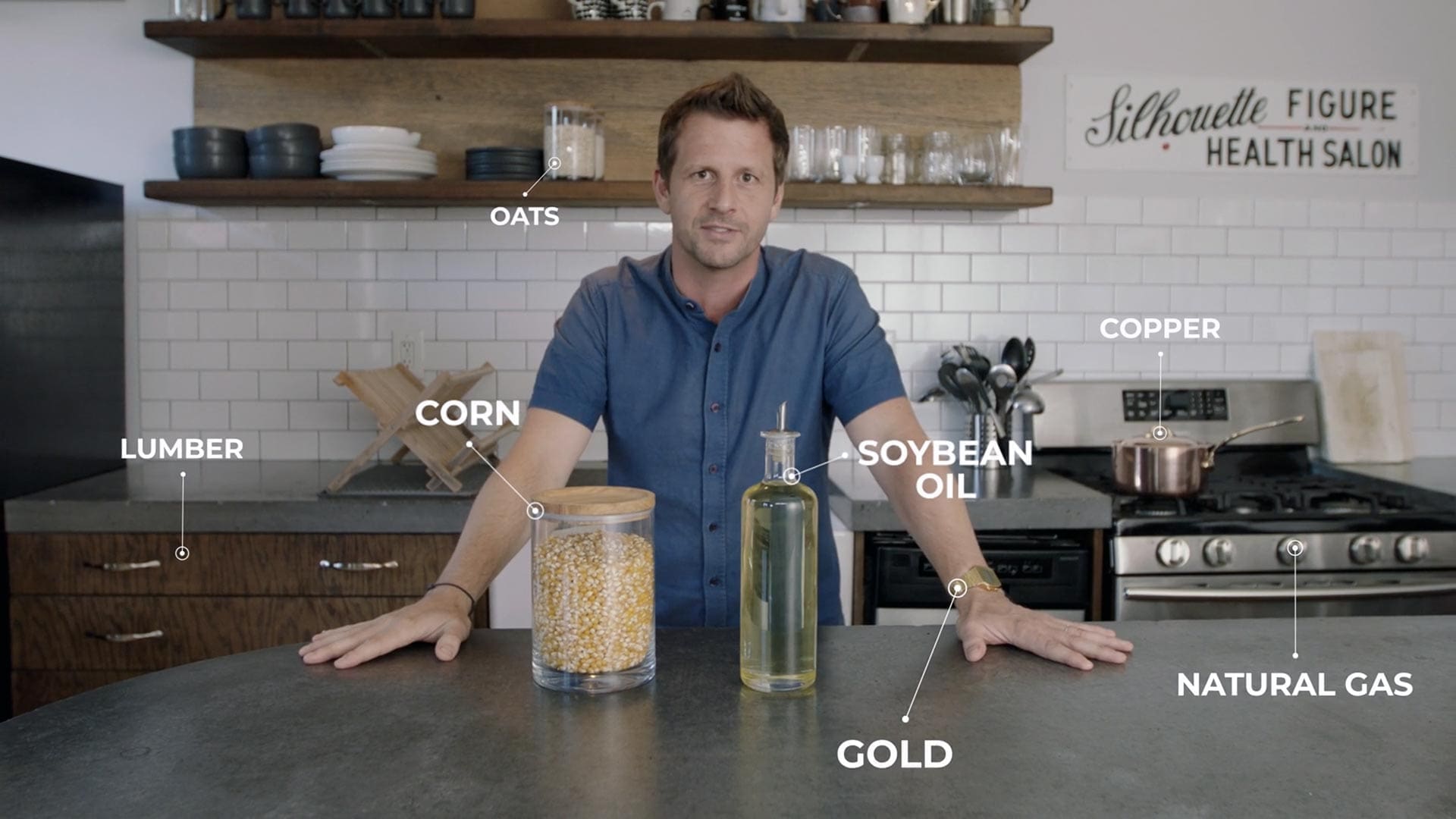 Jamie McDonald standing in a kitchen with labeled commodities including corn, oats, soybean oil, lumber, gold, copper, and natural gas, illustrating futures trading concepts.